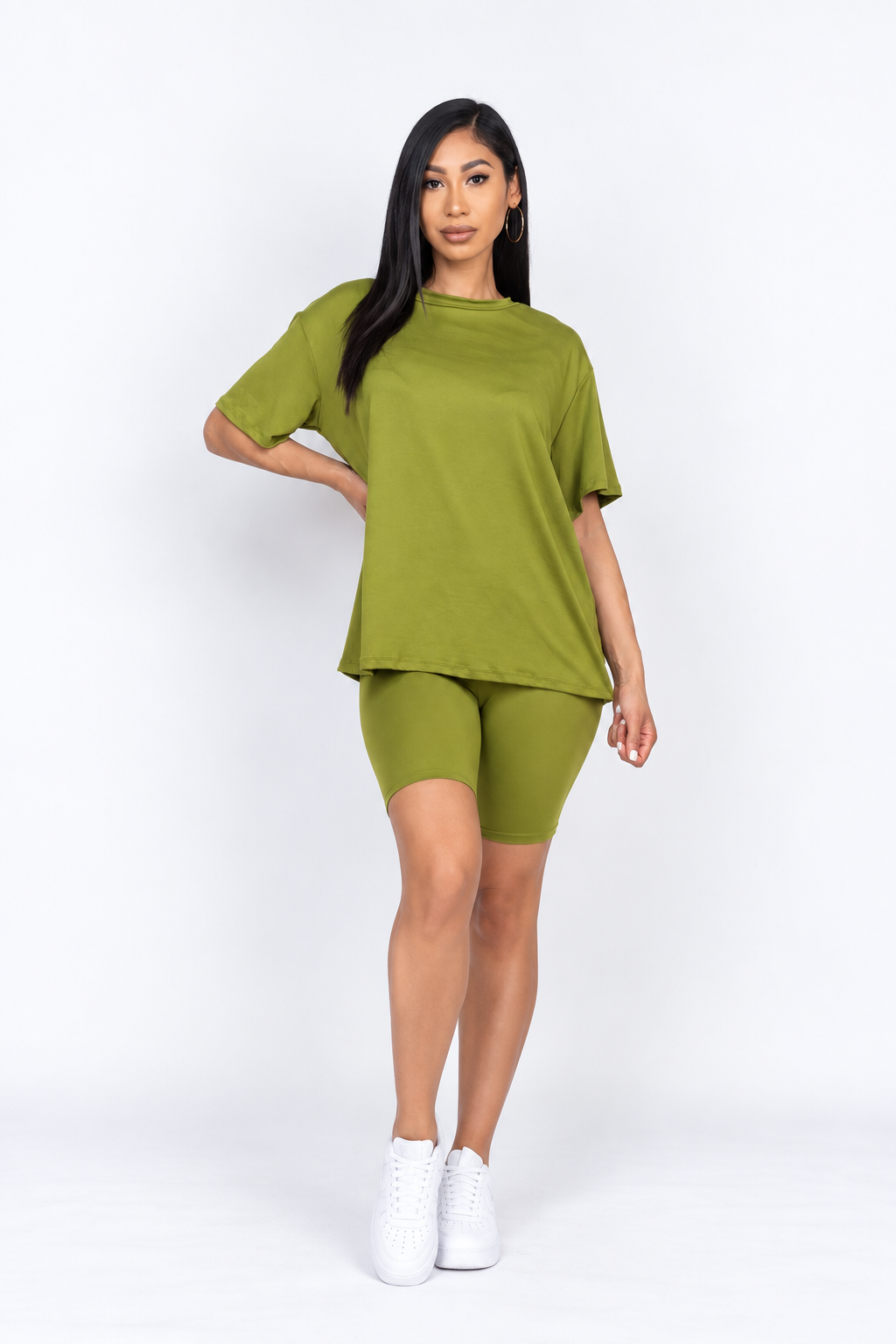 A woman modeling a matching olive green lounge set consisting of a short-sleeve crewneck t-shirt and high-waisted biker shorts, paired with white sneakers, against a plain white studio background.