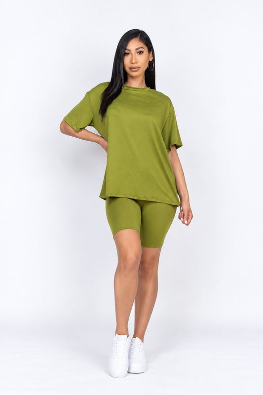 A woman modeling a matching olive green lounge set consisting of a short-sleeve crewneck t-shirt and high-waisted biker shorts, paired with white sneakers, against a plain white studio background.