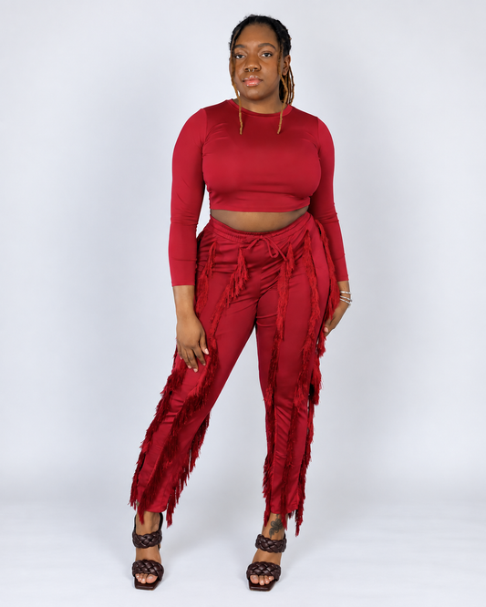 A woman modeling a matching red two-piece outfit consisting of a long-sleeve crop top and high-waisted fringed pants, paired with brown strappy heels, against a light gray studio background.