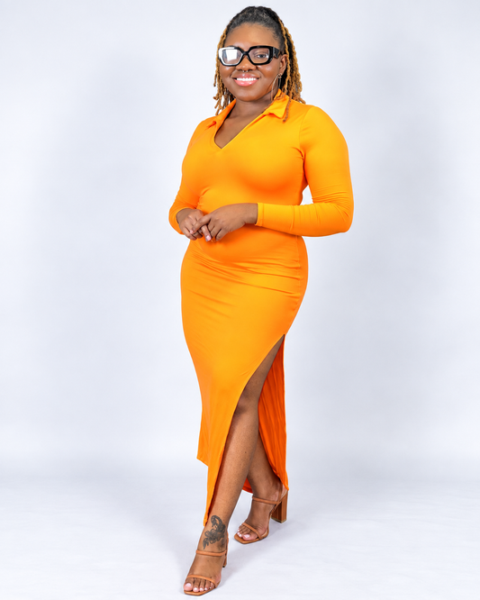 A woman modeling a bright orange long-sleeve collared maxi dress with a high side slit, paired with clear-framed glasses and brown heeled sandals, against a light gray studio background.