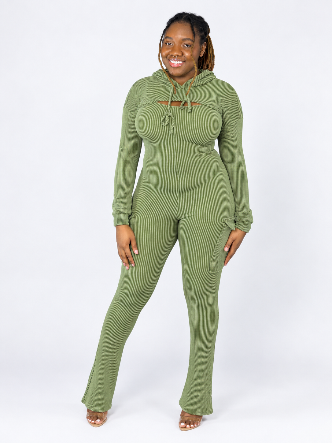 A woman modeling a sage green ribbed knit jumpsuit, drawstring hood, and side pockets, standing against a plain white studio background.