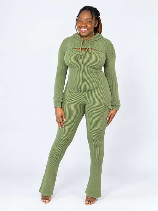 A woman modeling a sage green ribbed knit jumpsuit, drawstring hood, and side pockets, standing against a plain white studio background.