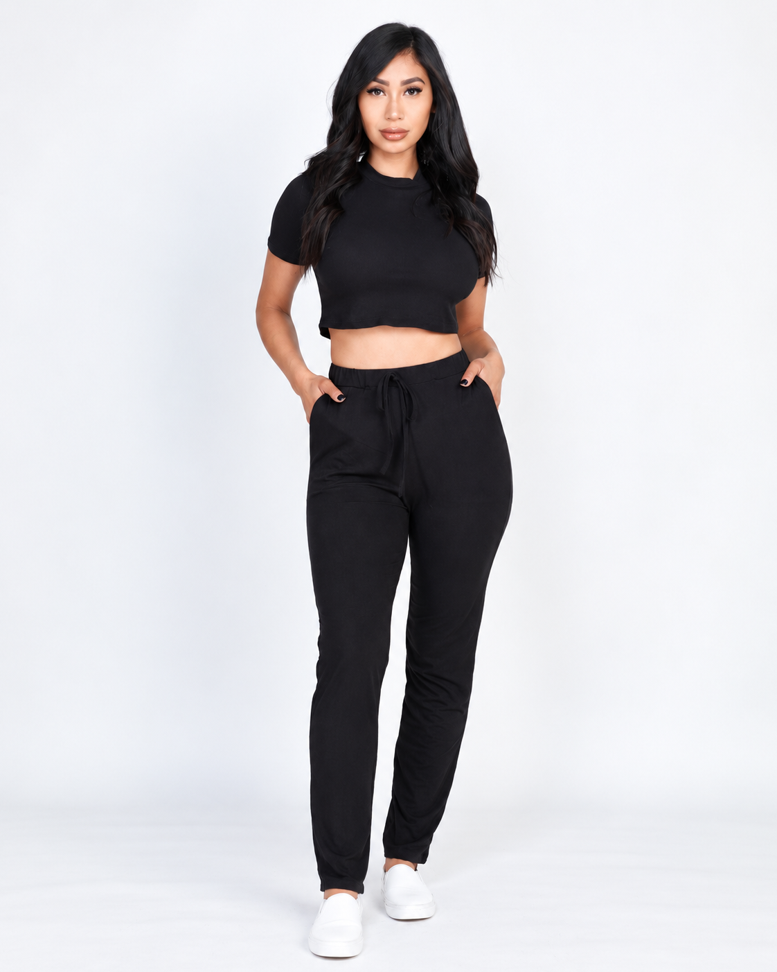 A woman modeling a black short-sleeve crop top and black faux drawstring pants against a plain white studio background.