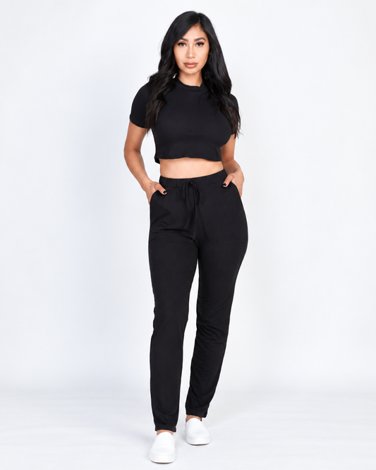 A woman modeling a black short-sleeve crop top and black faux drawstring pants against a plain white studio background.