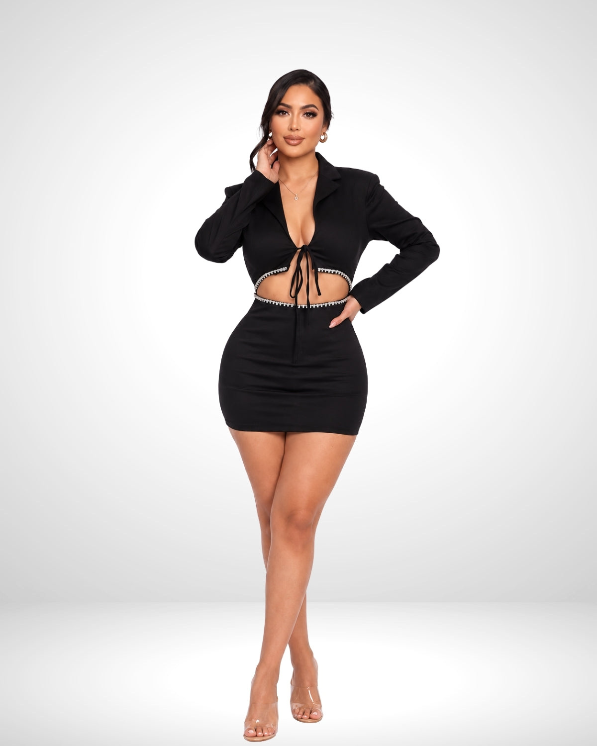 A woman modeling a black long-sleeve mini dress with a tie-front detail and cutout waist, paired with clear high-heeled sandals, against a light gray studio background.